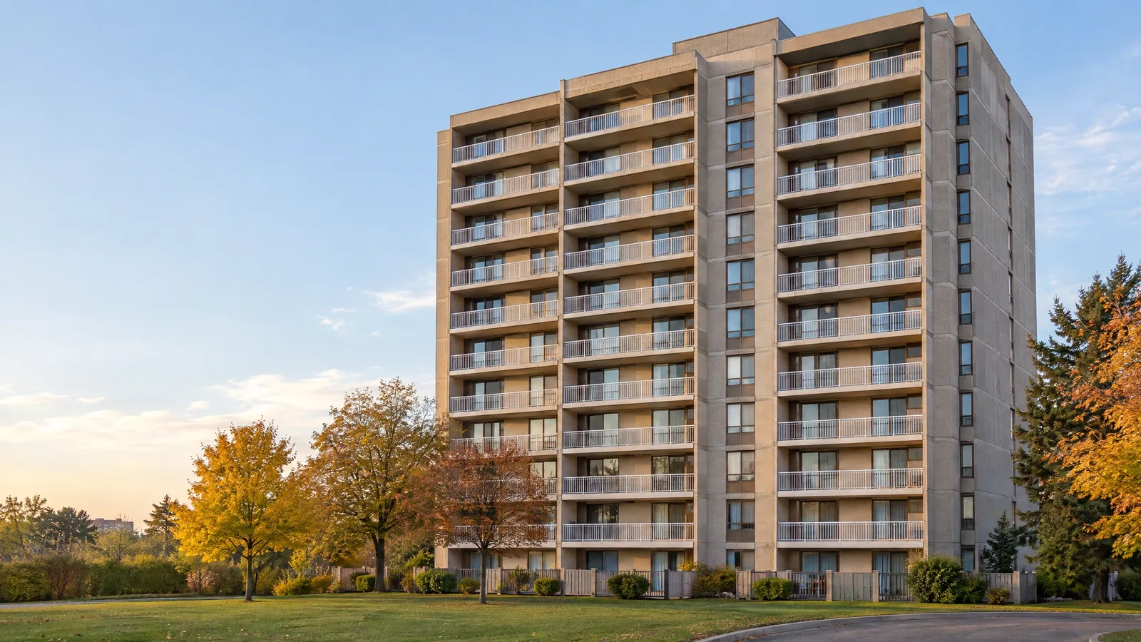 Concrete condo tower from the 1980s with stacked balconies and bronze aluminum sliding doors, autumn trees on the lawn in late-day light