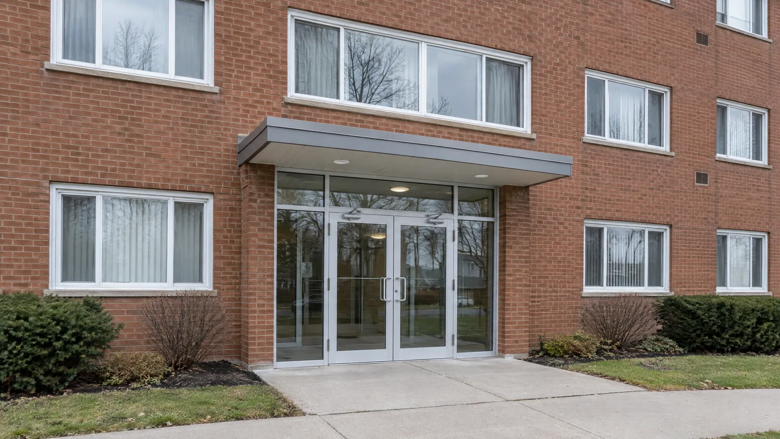 Brick walk-up apartment building with aluminum double glass entrance doors and glass sidelights under a flat metal canopy