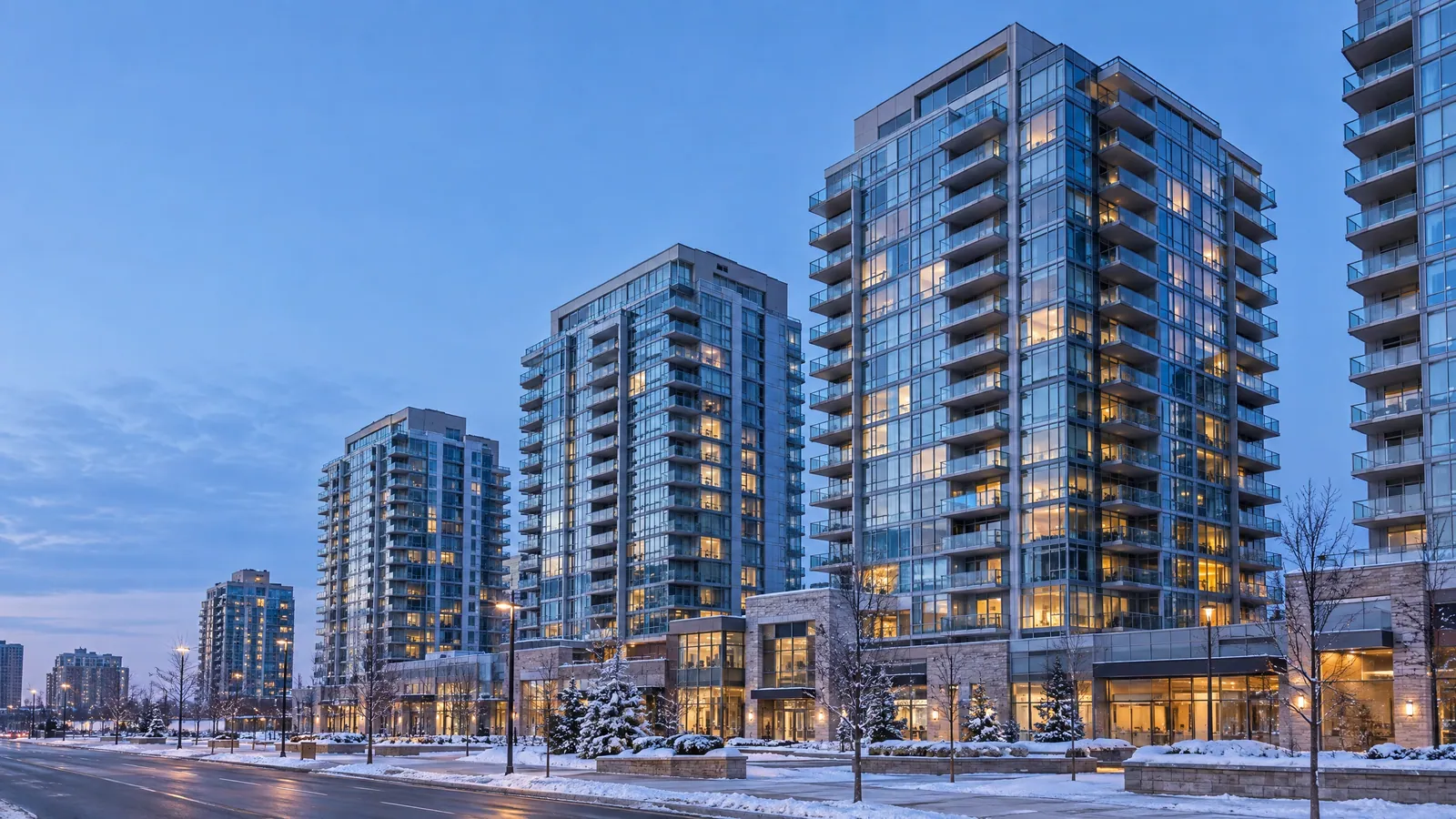Glass-clad condo towers with rows of balconies above a stone-faced podium and a snowy sidewalk at winter dusk