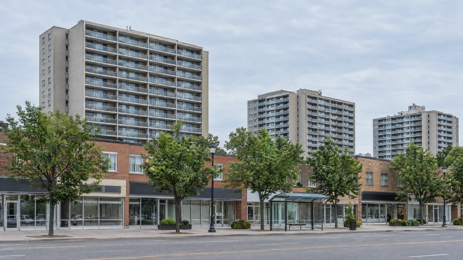 Three concrete apartment towers with rows of balconies rising behind a strip of two-storey brick shops with glass storefronts and a bus shelter in summer