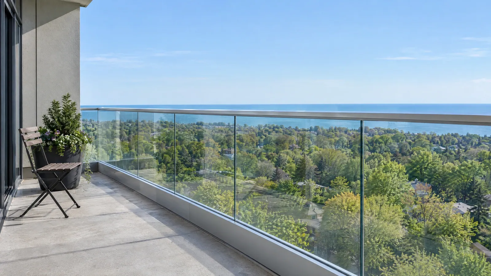 High condo balcony edged with clear glass guard panels in a metal base shoe under a top rail, looking over treetops to the lake