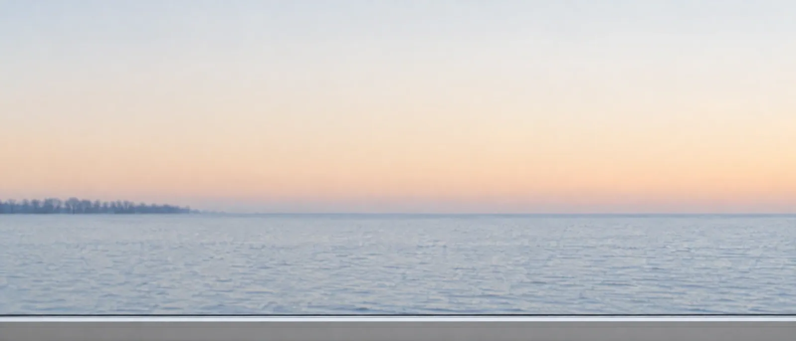 Wide, calm view of a pale lake horizon at winter dawn, seen past a white window frame