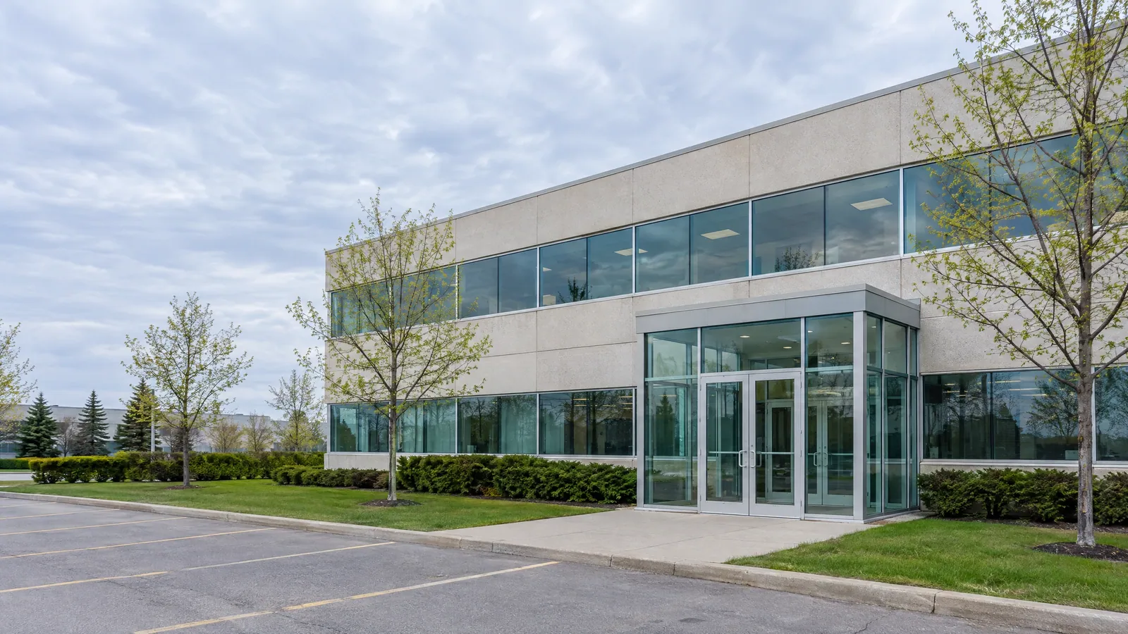 Two-storey suburban office with bands of ribbon windows and a glass entrance vestibule beside a parking lot in spring