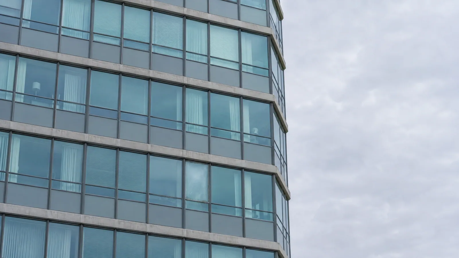 Looking up at a residential tower's glass window-wall under a grey sky, with one hazy failed unit among the clear ones and slab edges between floors
